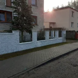 Nowoczesny, jasnoszary mur z bloczków betonowych, tworzący ogrodzenie posesji z elementami ażurowymi, widoczny z perspektywy ulicy z chodnikiem z kostki brukowej i fragmentem drogi gruntowej.