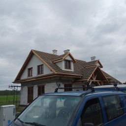 Zakład Ogólnobudowlany ŁUK-BUD Łukasz Wróblewski - Budowa domu jednorodzinnego w trakcie realizacji, widoczne ściany z bloczków, drewniana konstrukcja dachu, częściowo położona dachówka, na pierwszym planie niebieski samochód dostawczy.