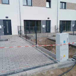 Nowe szeregowe domy z szarą kostką brukową na podjeździe, ogrodzone siatką, z elementami instalacji elektrycznej na zewnątrz.
