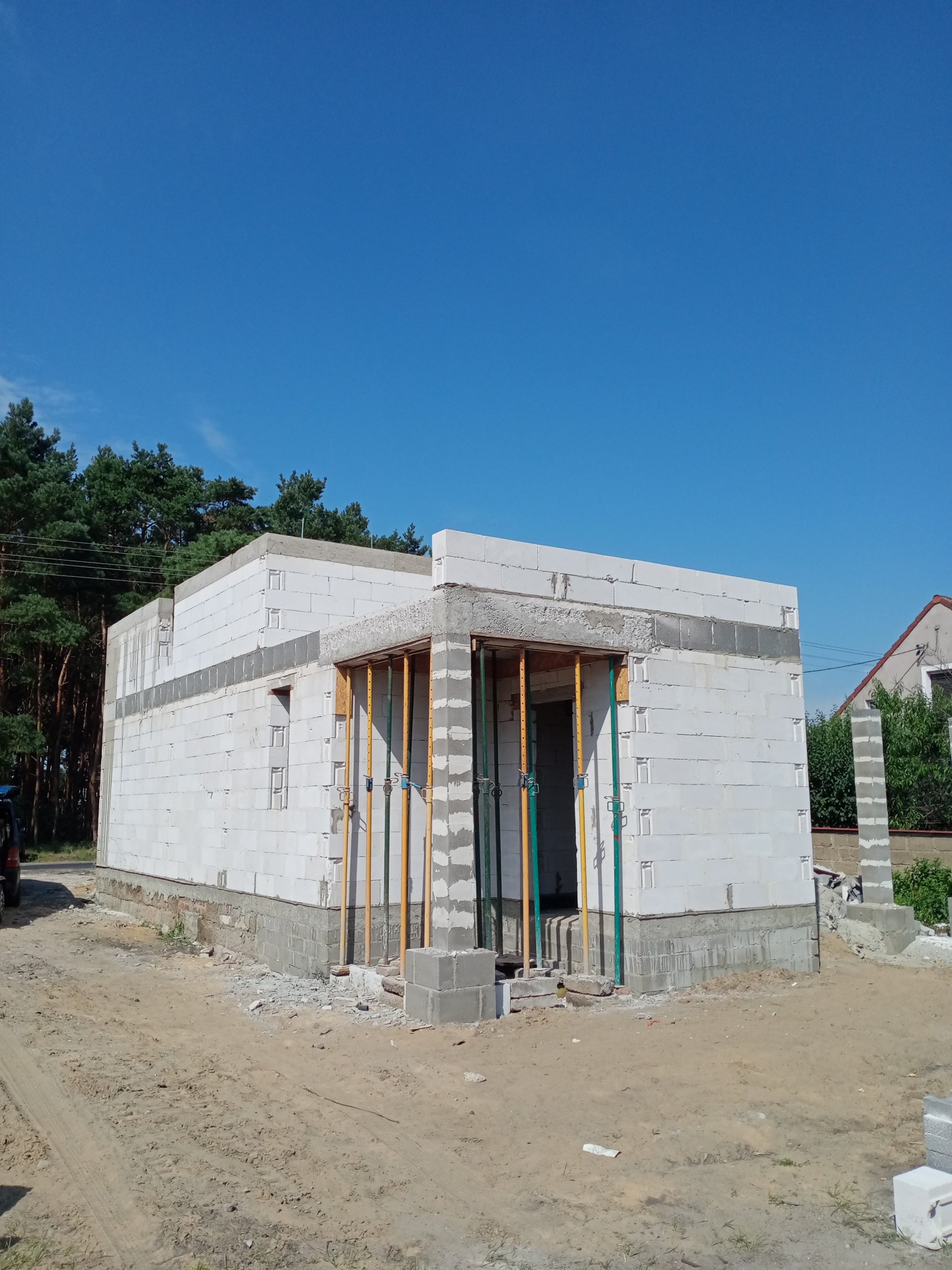 Wznoszenie ścian parterowego domu z białych bloczków, widoczne podpory szalunkowe, fragment fundamentu oraz teren budowy w słoneczny dzień.