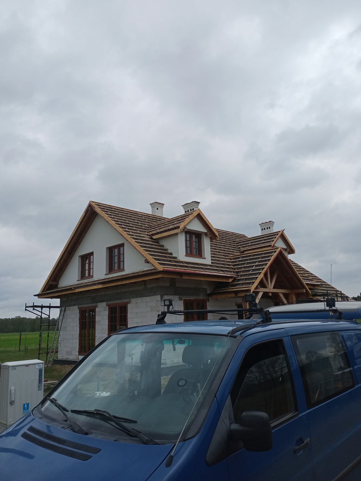 Budowa domu jednorodzinnego w trakcie realizacji, widoczne ściany z bloczków, drewniana konstrukcja dachu, częściowo położona dachówka, na pierwszym planie niebieski samochód dostawczy.