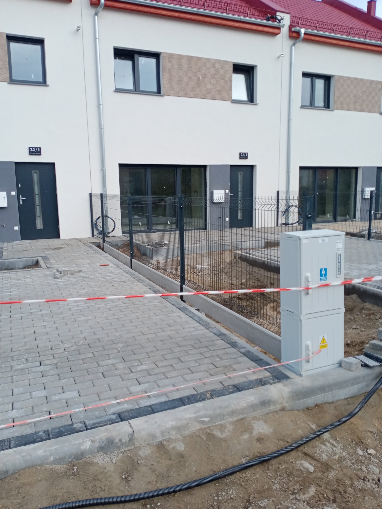 Nowe szeregowe domy z szarą kostką brukową na podjeździe, ogrodzone siatką, z elementami instalacji elektrycznej na zewnątrz.