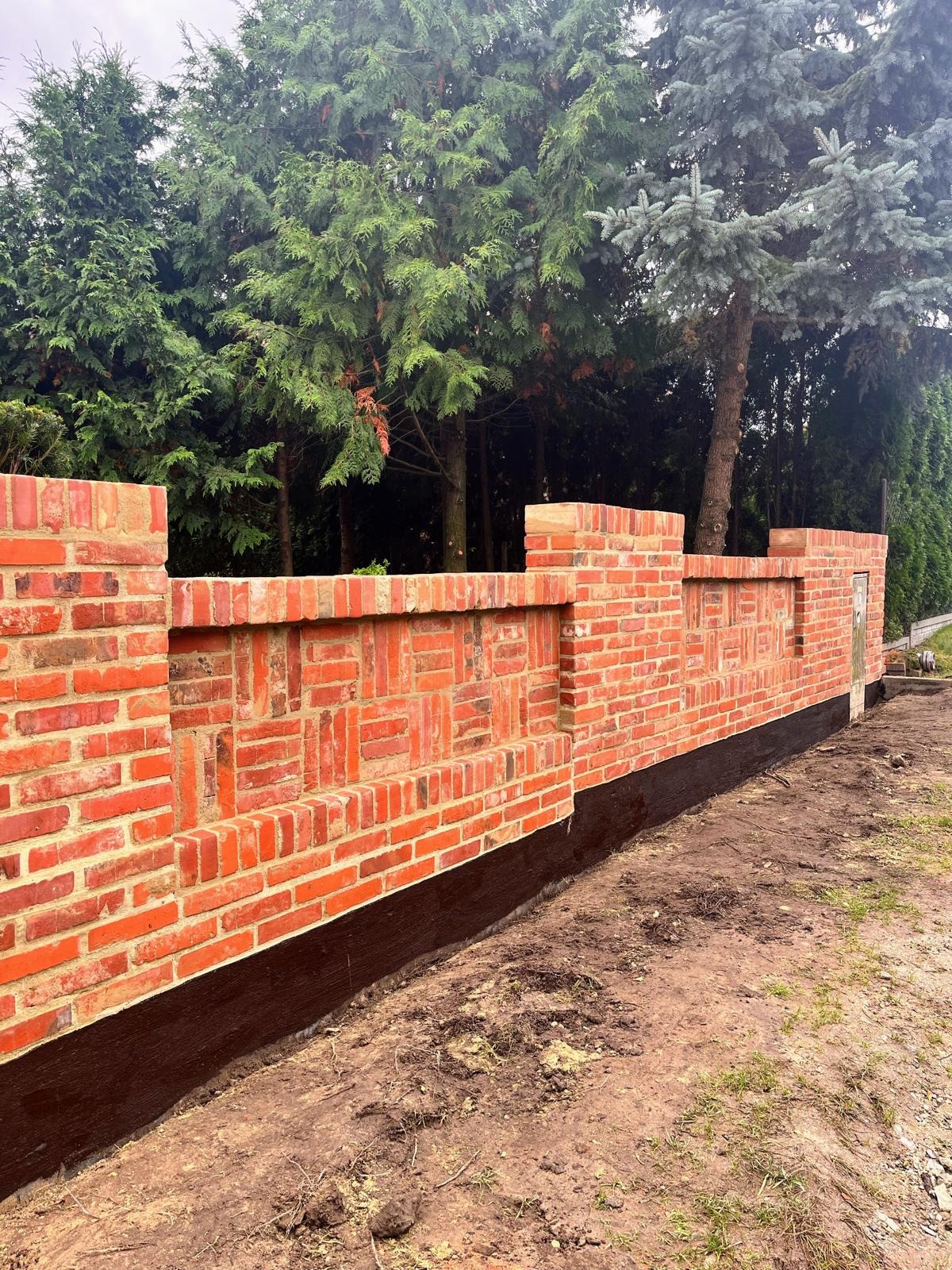 Nowo wymurowany, niski murek z czerwonej cegły, z dekoracyjnym wzorem ułożenia cegieł, zabezpieczony czarną izolacją przy gruncie, na tle zieleni.