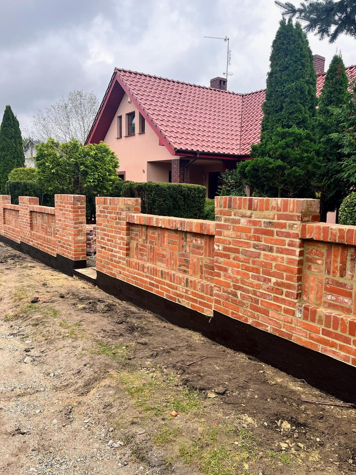 Nowo wymurowany, dekoracyjny mur z czerwonej cegły na fundamencie z izolacją bitumiczną, na tle domu z czerwoną dachówką i zieleni. Widok z poziomu gruntu.