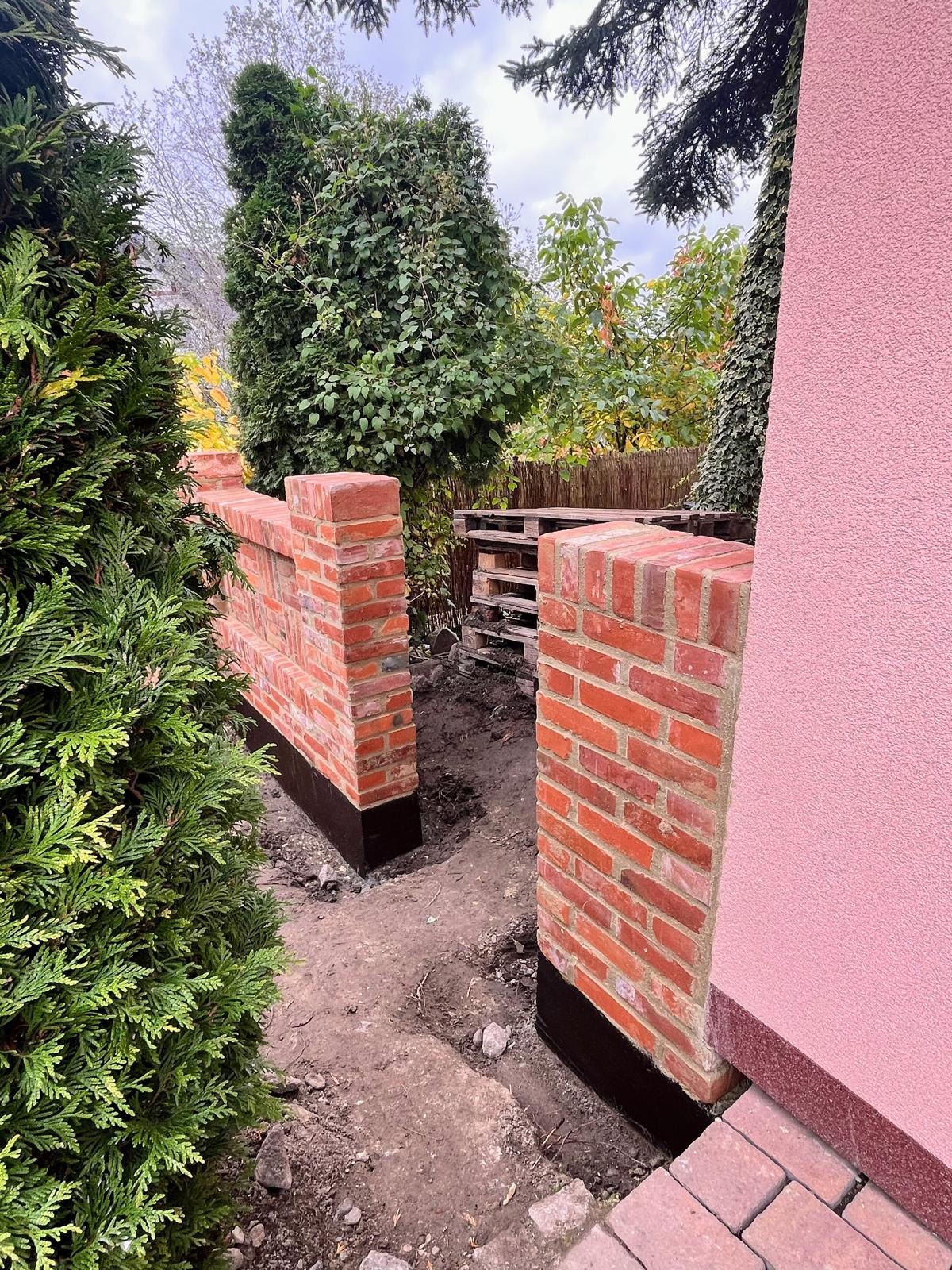 Ceglana ściana w trakcie budowy, z widocznym zabezpieczeniem przeciwwilgociowym, sąsiadująca z różową elewacją domu i zielenią. Widoczne palety w tle.