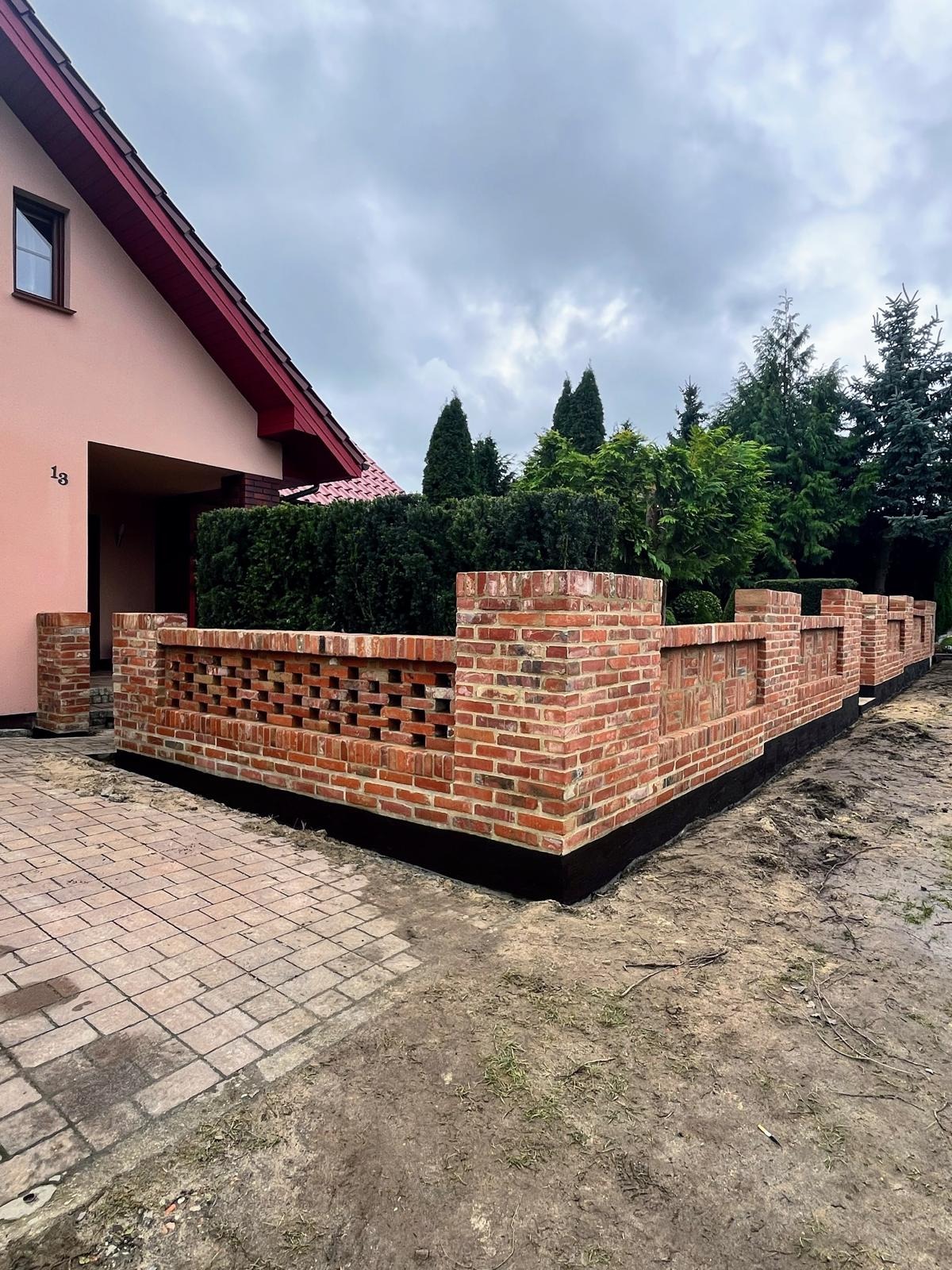Nowo wybudowany, dekoracyjny mur z czerwonej cegły, oddzielający posesję od ulicy. Widoczny fragment elewacji domu z numerem 13. Tło stanowią drzewa i pochmurne niebo.