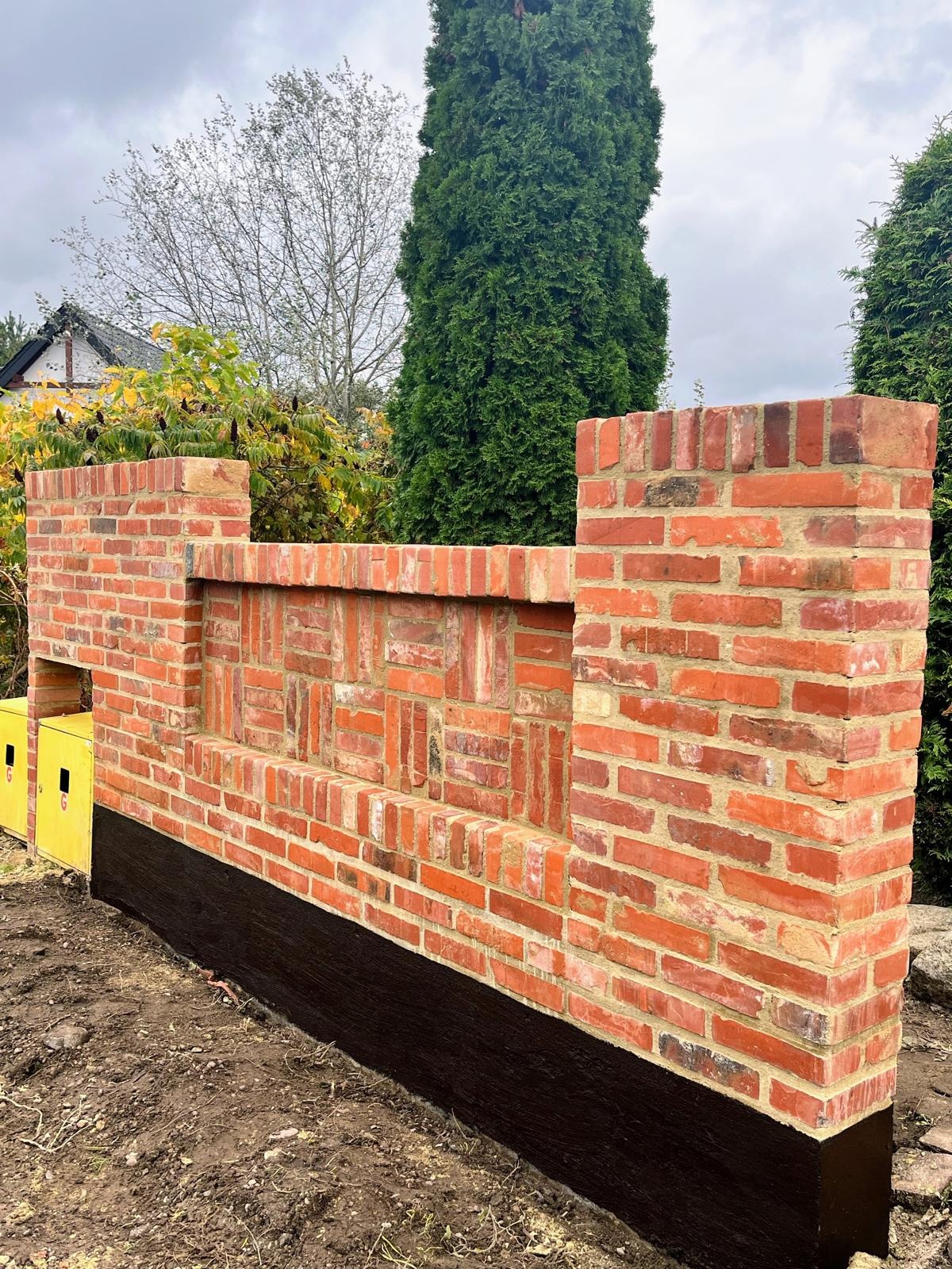 Nowo wymurowany, dekoracyjny murek z czerwonej cegły z czarną izolacją, otoczony zielenią. Widoczna faktura cegieł i wzór ułożenia. Element architektury krajobrazu w ogrodzie.