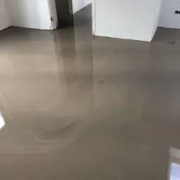 Beżowa, błyszcząca podłoga w nowym, pustym pomieszczeniu z białymi ścianami, widoczne odbicia okien, gładka powierzchnia bez spoin.