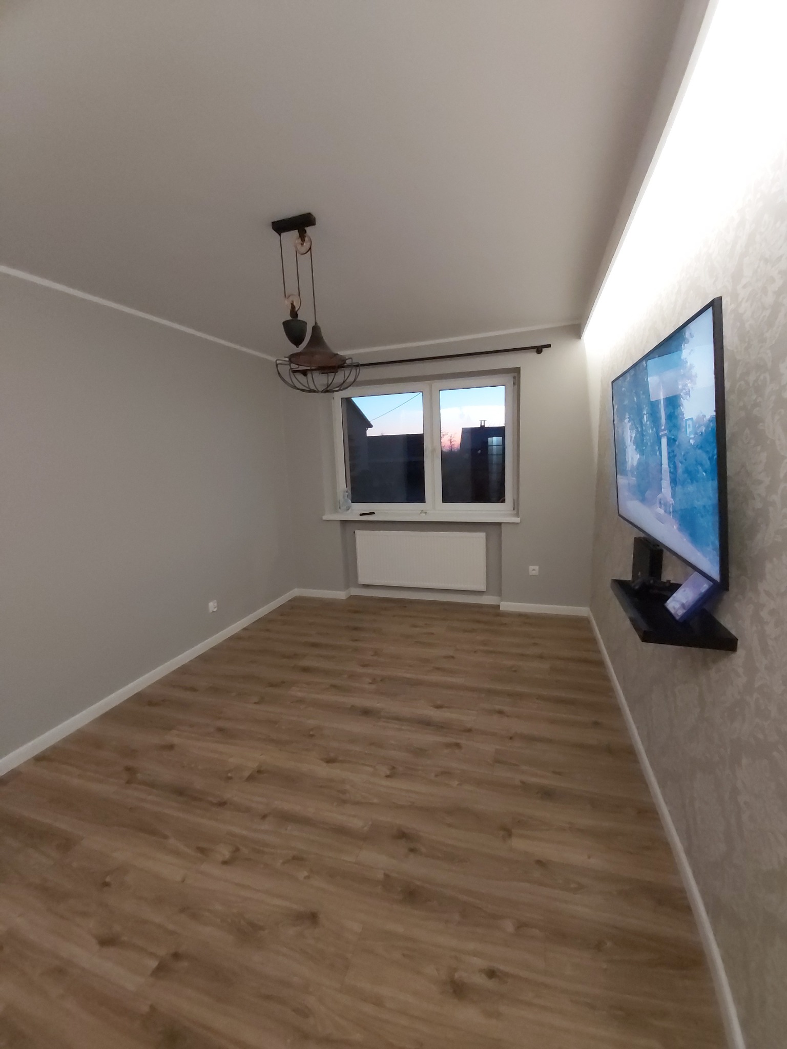 Wykończony pokój w Spławiu z drewnianą podłogą, szarą ścianą i telewizorem na ozdobnej tapecie, oświetlony lampą w stylu industrialnym.