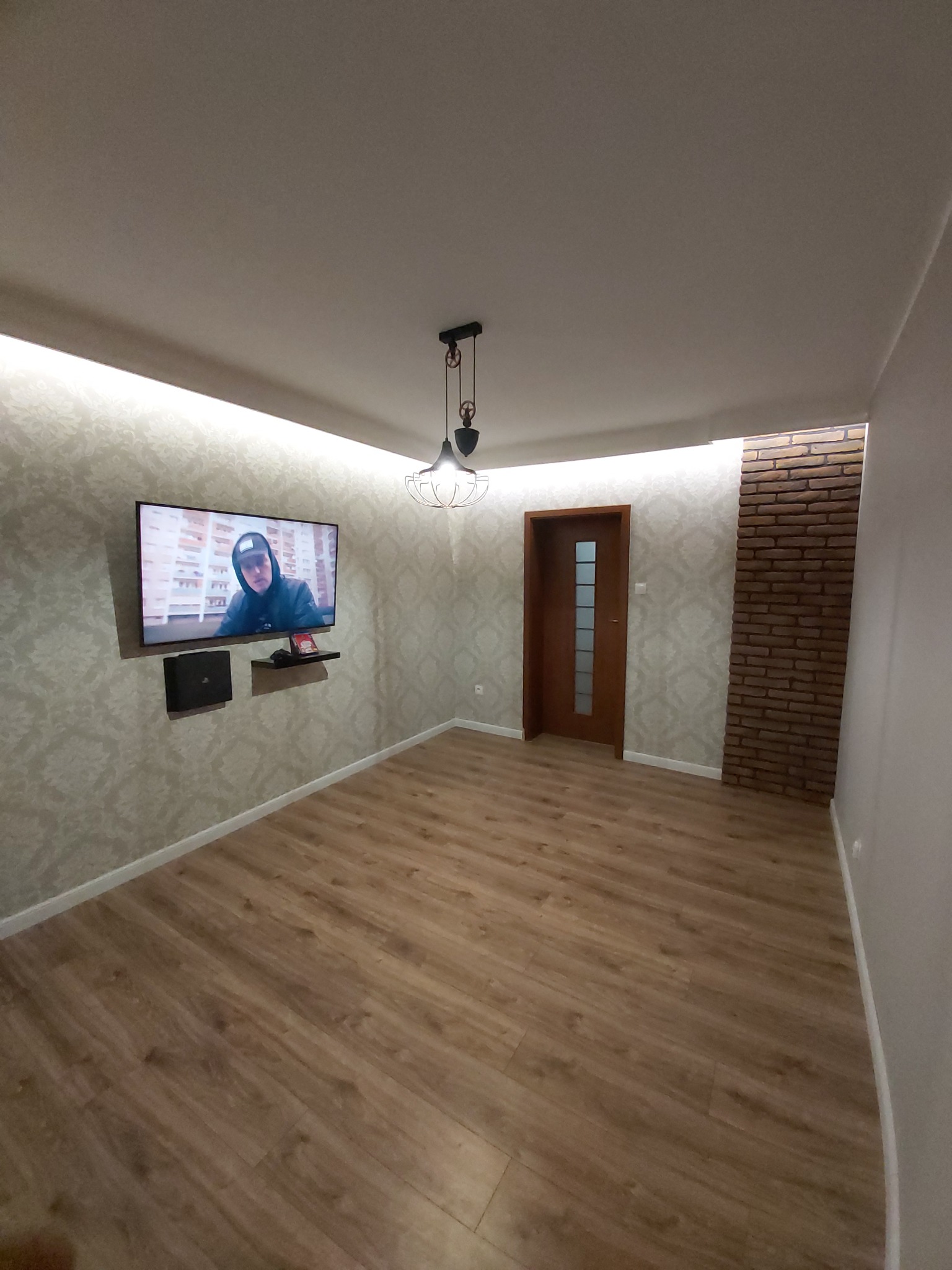 Pokój po remoncie z panelami na podłodze, tapetą w ornamenty, fragmentem ściany z cegły dekoracyjnej i podwieszanym sufitem z oświetleniem LED, telewizor na ścianie.