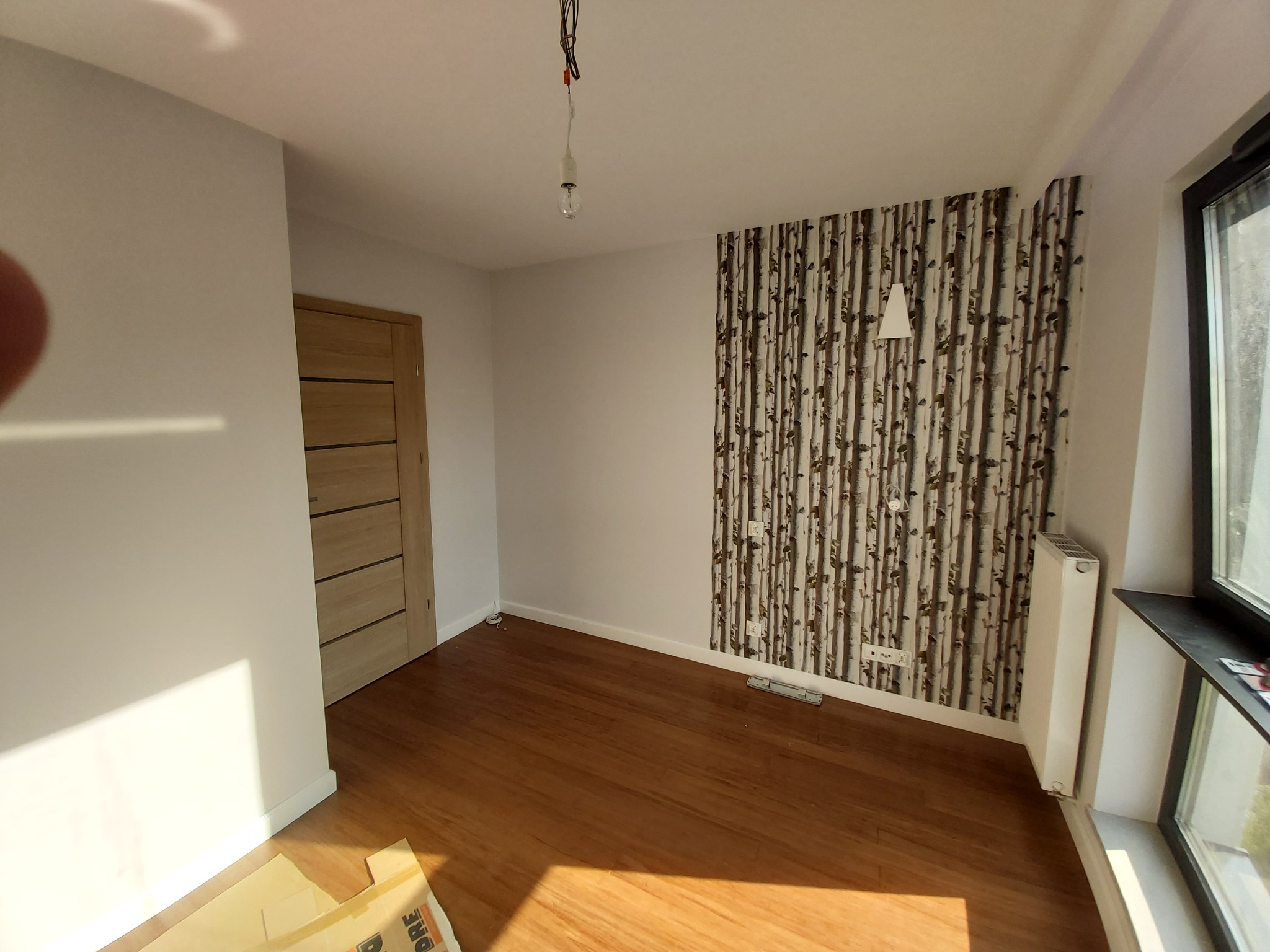 Pokój po odświeżeniu: drewnopodobna podłoga, ściana z tapetą imitującą brzozowy las, białe ściany, drewniane drzwi, grzejnik przy oknie, surowa żarówka zwisająca z sufitu.