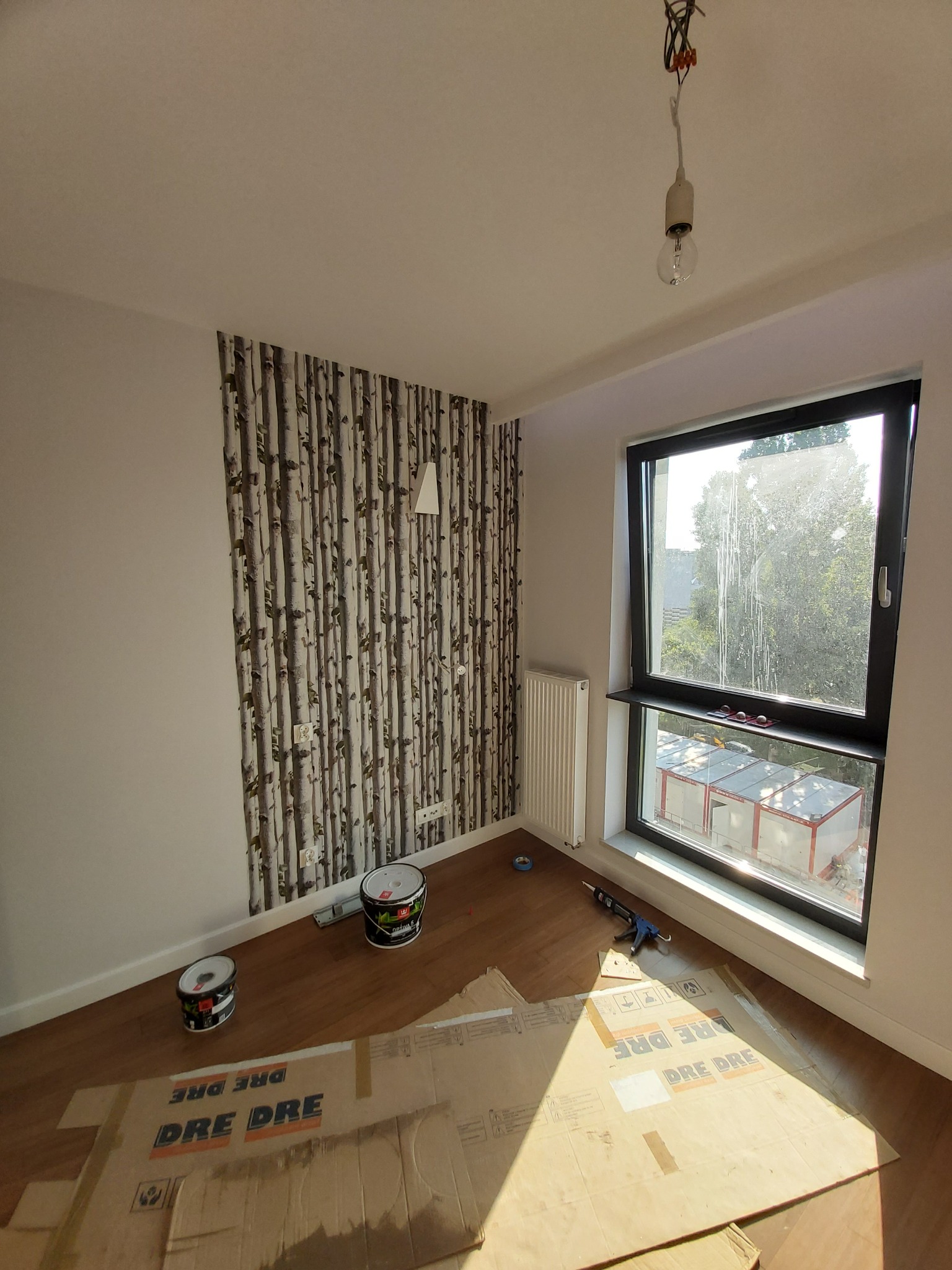 Pokój w trakcie prac wykończeniowych: ściana z tapetą w motywie brzozowego lasu, okno z czarną ramą, grzejnik, dwie puszki farby, kartony zabezpieczające podłogę, wisząca żarówka bez klosza.