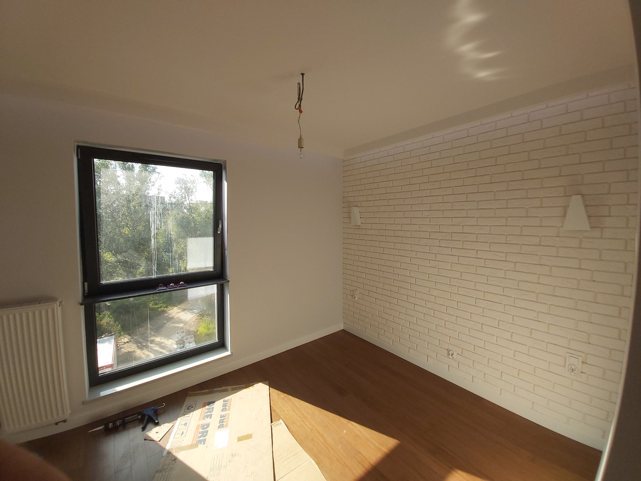 Wnętrze pokoju po remoncie: ściana z cegły dekoracyjnej, drewniana podłoga, okno z widokiem na zieleń, grzejnik, narzędzia i karton na podłodze.