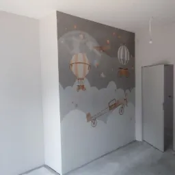 Pokój po odświeżeniu, z jedną ścianą ozdobioną szarą tapetą z motywem balonów, samolotów i chmur w stylu dziecięcym, pozostałe ściany pomalowane na biało, widoczne gniazdka elektryczne...