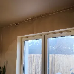 Świeżo nałożona, mokra gładź na ścianach i suficie w pokoju z oknem, widoczne ślady po szpachlowaniu przy listwie sufitowej.