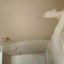 Gładzie i Malowanie Natryskowe - Świeżo położona, mokra warstwa gładzi na suficie w trakcie prac remontowych, widoczne lampy sufitowe i fragmenty mebli zabezpieczonych folią.