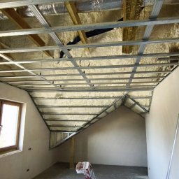 Gładzie i Malowanie Natryskowe - Poddasze z oknem w trakcie izolacji pianą i montażu stelażu sufitu podwieszanego, drabina w pomieszczeniu. Widok na skośny sufit i ścianę.