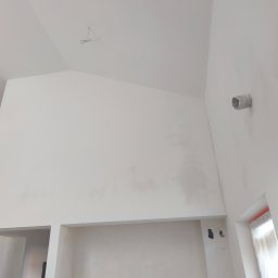 Gładzie i Malowanie Natryskowe - Wnętrze po szpachlowaniu i malowaniu na biało. Widoczna wysoka ściana, sufit ze skosem oraz niedokończone gniazdka elektryczne. Na ścianie widoczny przewód wentylacyjny.