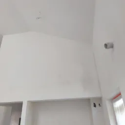 Wnętrze po szpachlowaniu i malowaniu na biało. Widoczna wysoka ściana, sufit ze skosem oraz niedokończone gniazdka elektryczne. Na ścianie widoczny przewód wentylacyjny.