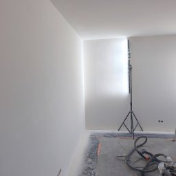 Gładzie i Malowanie Natryskowe - Wnętrze po szpachlowaniu: gładkie, białe ściany podświetlone lampą budowlaną. Na podłodze widoczny sprzęt do szlifowania i zabezpieczenia.