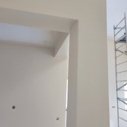 Gładzie i Malowanie Natryskowe - Świeżo pomalowane na beżowo ściany z widocznymi otworami na gniazdka i fragment rusztowania, ukazujące etap wykończeniowy w jasnym wnętrzu.