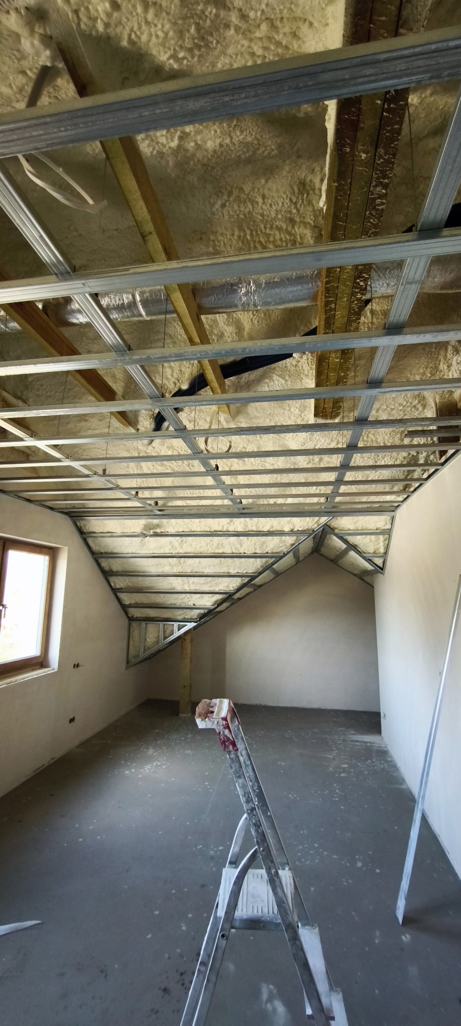 Poddasze z oknem w trakcie izolacji pianą i montażu stelażu sufitu podwieszanego, drabina w pomieszczeniu. Widok na skośny sufit i ścianę.