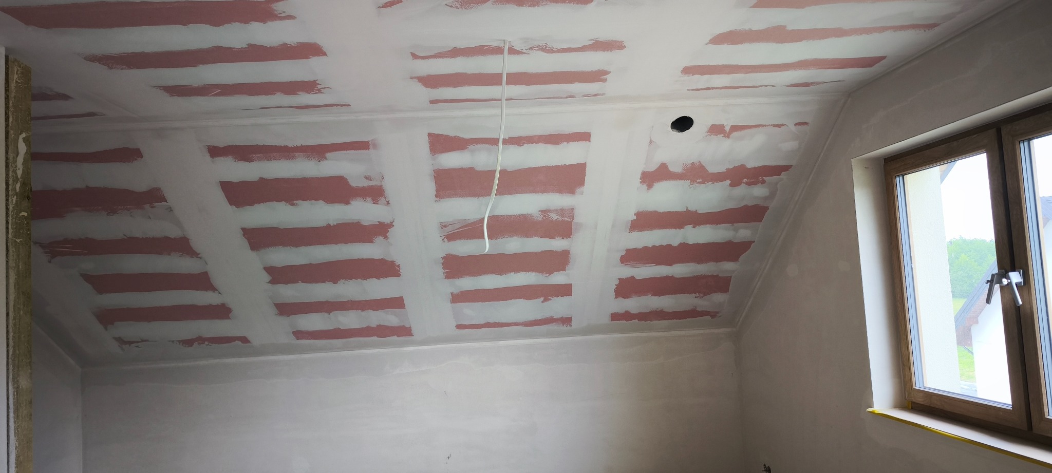Skośny sufit poddasza po szpachlowaniu, z widocznymi fragmentami różowego podkładu, oczekujący na dalsze prace wykończeniowe. Widoczne okno i kabel.