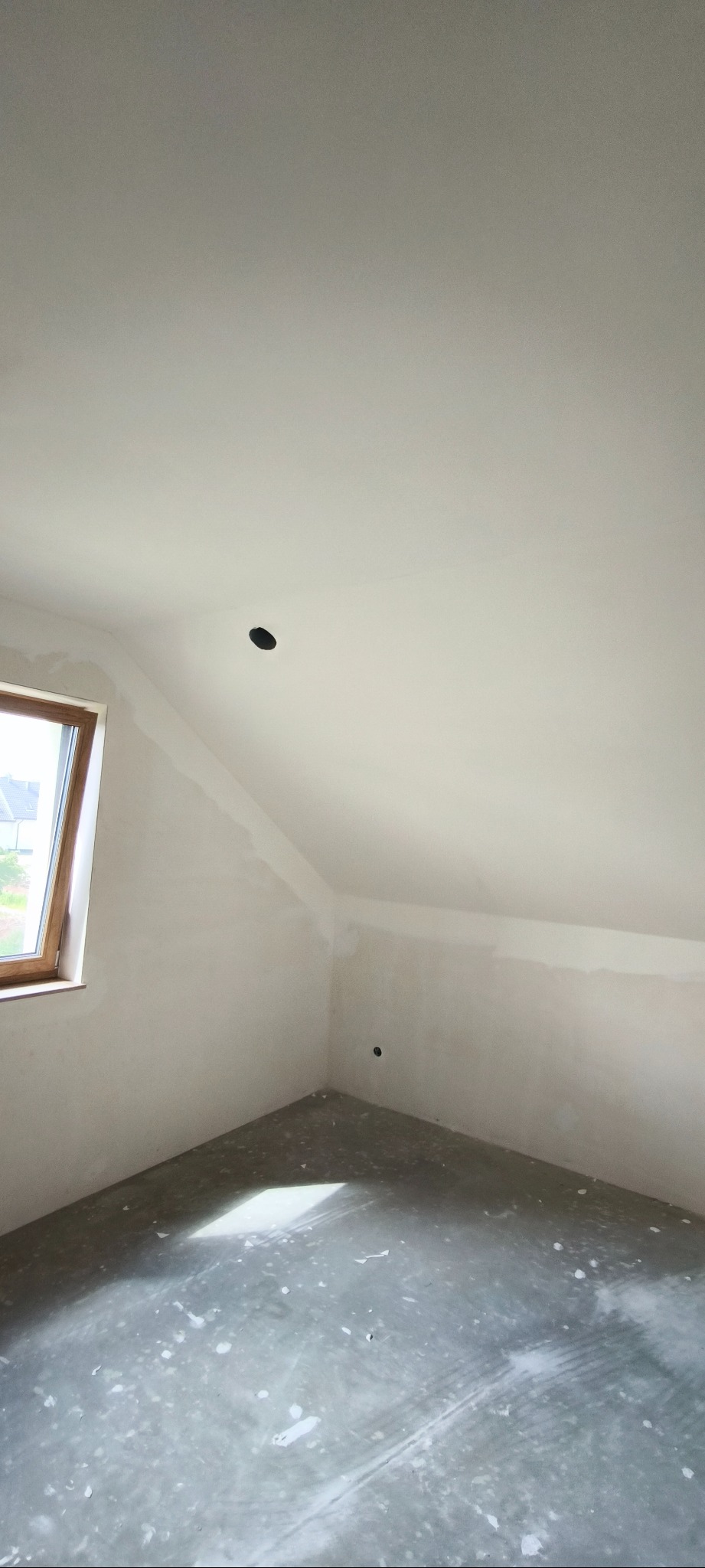 Poddasze po szpachlowaniu, z oknem i widocznymi punktami oświetleniowymi. Ściany i sufit przygotowane do dalszych prac wykończeniowych, podłoga zabezpieczona.
