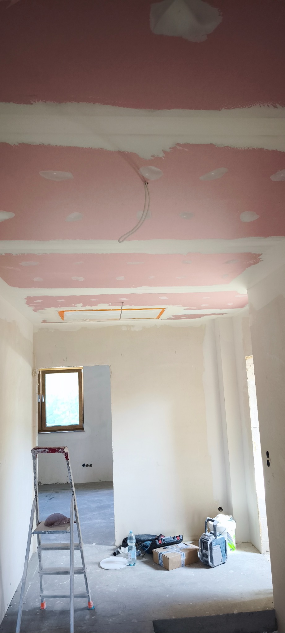 Wnętrze w trakcie prac wykończeniowych: ściany i sufit pokryte różową warstwą, widoczne narzędzia, drabina, okno. Częściowo zaszpachlowany sufit z instalacją.