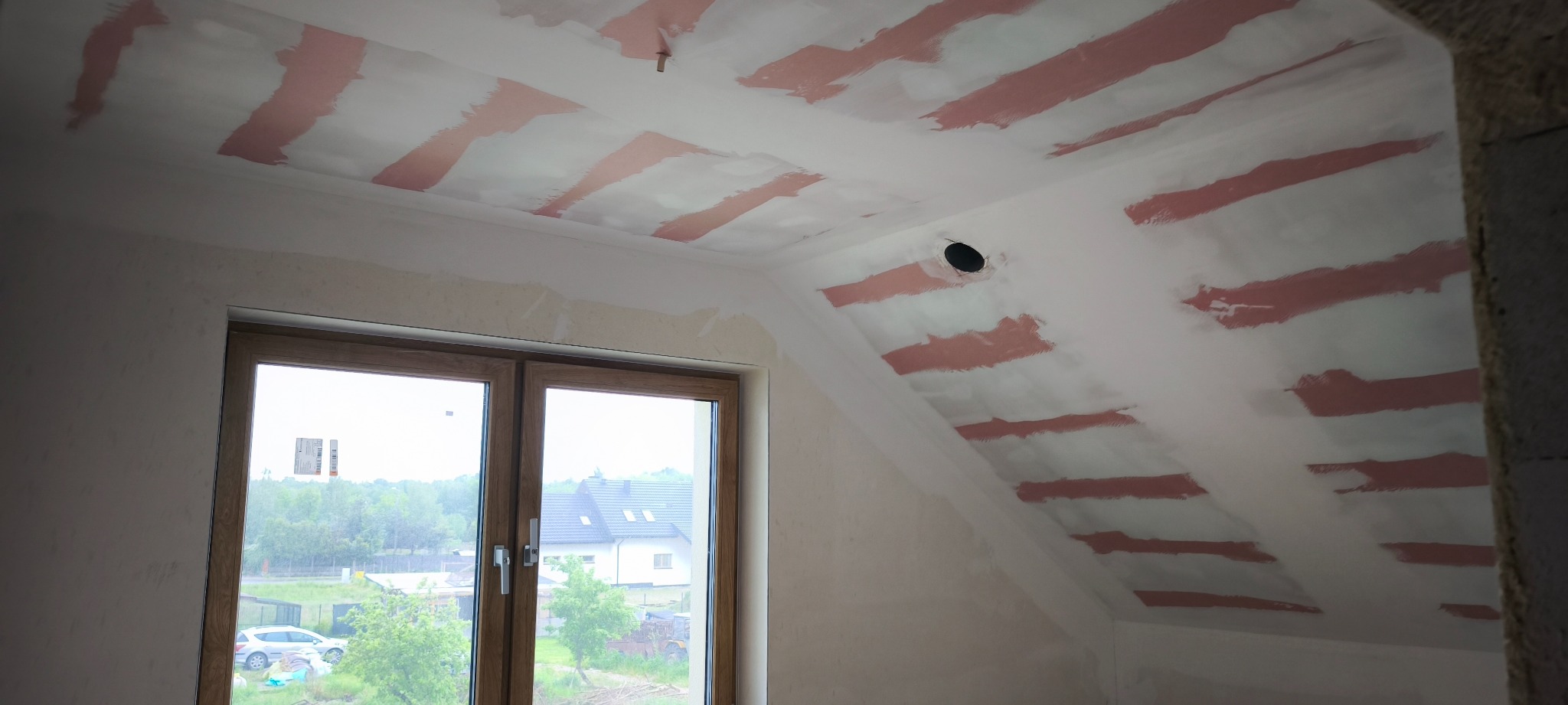 Poddasze w trakcie prac wykończeniowych: ściany i skośny sufit z pasami świeżo nałożonej gładzi szpachlowej. Widoczne okno i fragment ściany w surowym stanie.