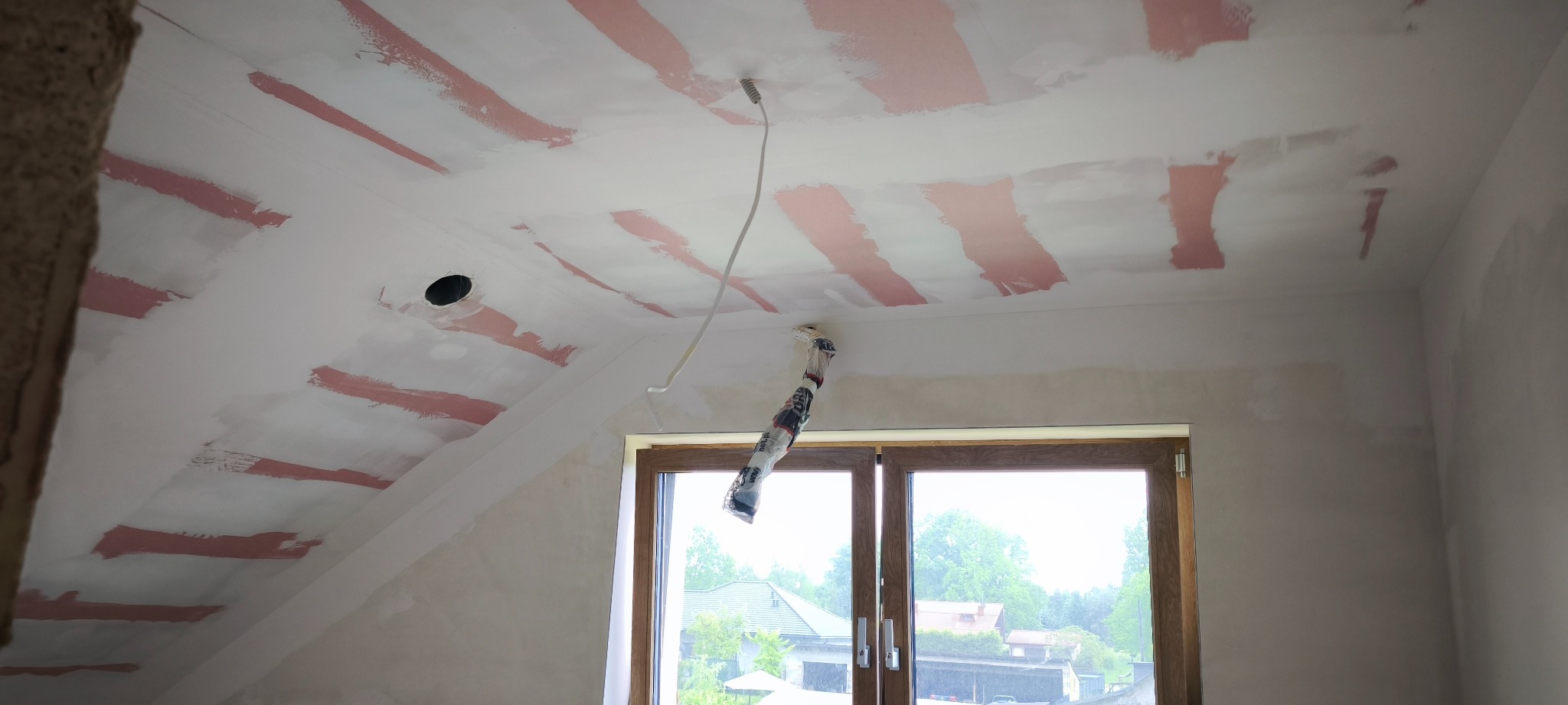 Skośne poddasze w trakcie prac przygotowawczych: widoczne fragmenty zagruntowanego sufitu w kolorze różowym, okno i wystający kabel elektryczny.