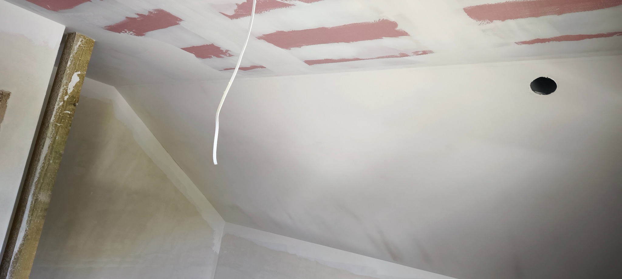 Sufit poddasza po szpachlowaniu z widocznymi łatami z różową farbą gruntującą, przygotowany do malowania. Widoczny otwór na oświetlenie i kabel.