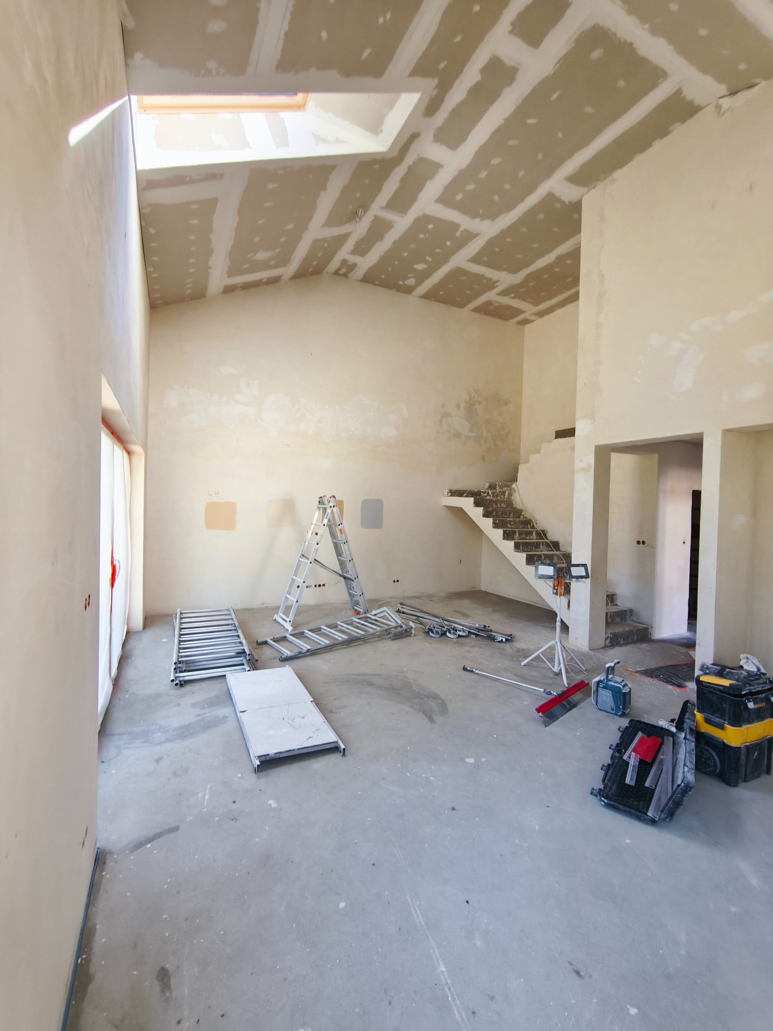 Wnętrze domu w trakcie prac wykończeniowych: ściany i sufit przygotowane do malowania, widoczne narzędzia i drabiny. Skosy i okno dachowe dodają charakteru.