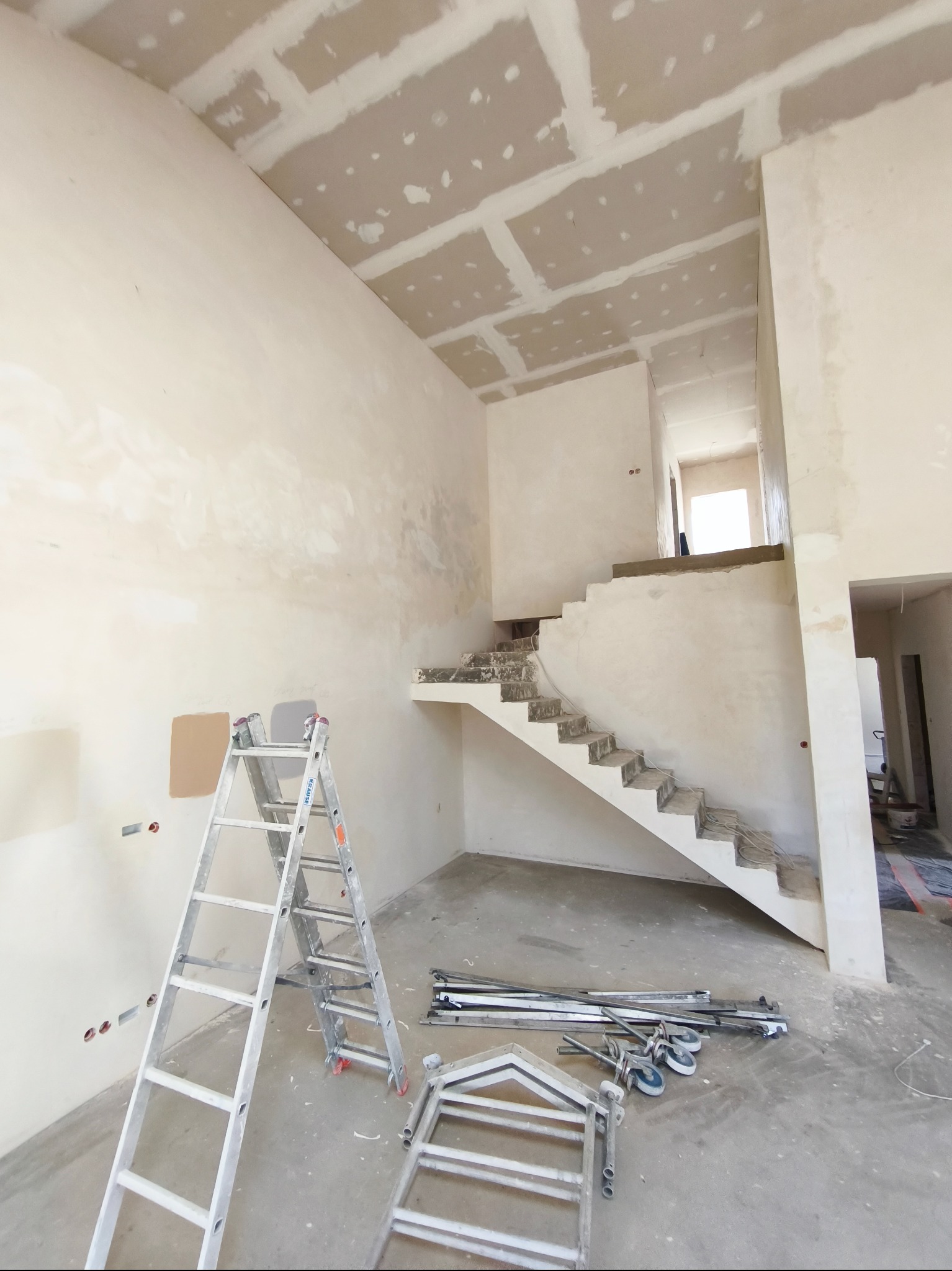 Wnętrze w trakcie prac wykończeniowych: ściany i sufit po szpachlowaniu, widoczne schody w stanie surowym, drabina malarska i narzędzia na podłodze.