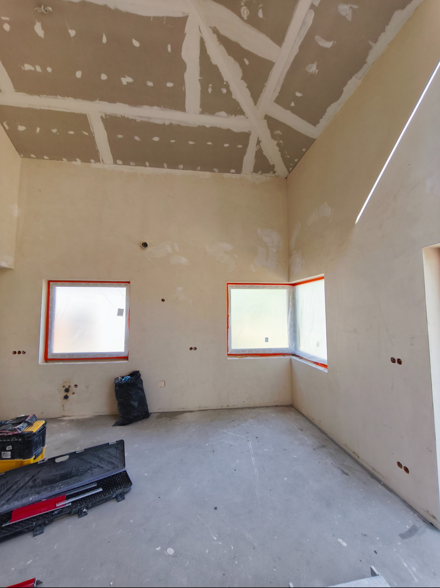 Wnętrze w trakcie szpachlowania: ściany i sufit częściowo pokryte gładzią, okna zabezpieczone folią, widoczne narzędzia i worki na gruz. Stan surowy.