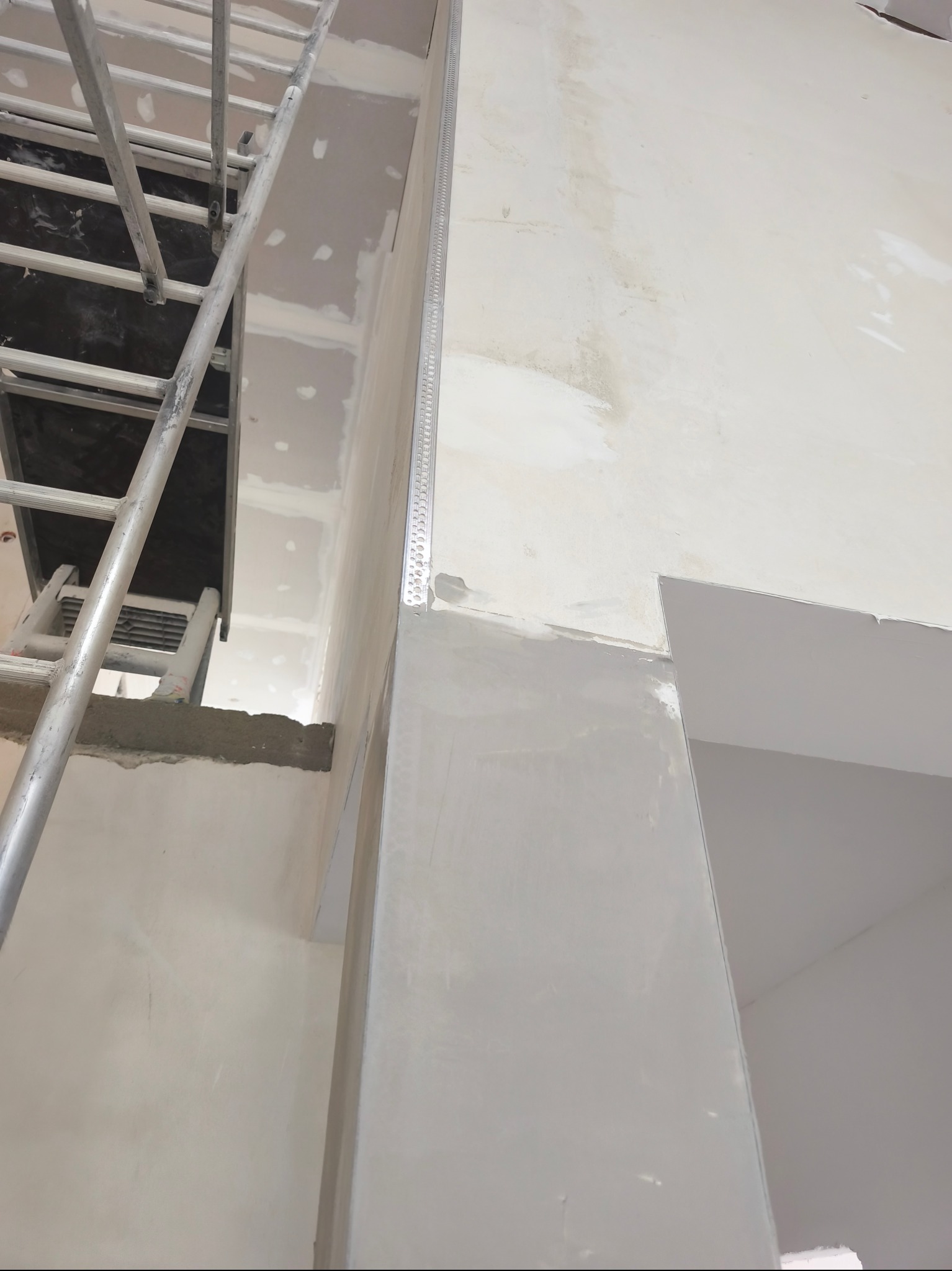 Fragment wnętrza w trakcie prac wykończeniowych: ściana z nałożoną gładzią, zamontowany narożnik aluminiowy i fragment rusztowania. Widoczne ślady szpachlowania na ścianie.