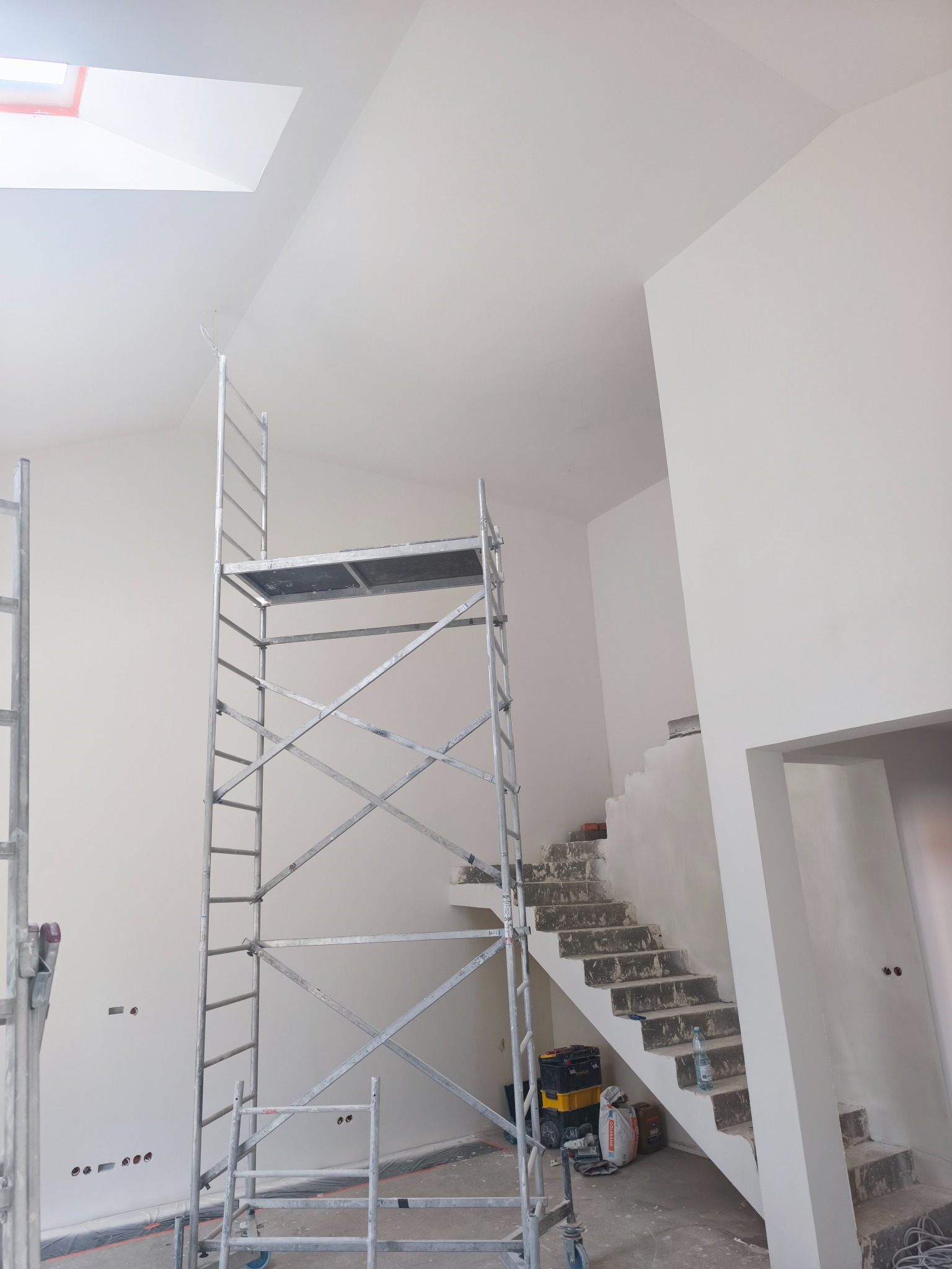 Wnętrze w trakcie prac wykończeniowych: białe ściany, schody w stanie surowym, rusztowanie. Widoczne narzędzia i materiały budowlane.