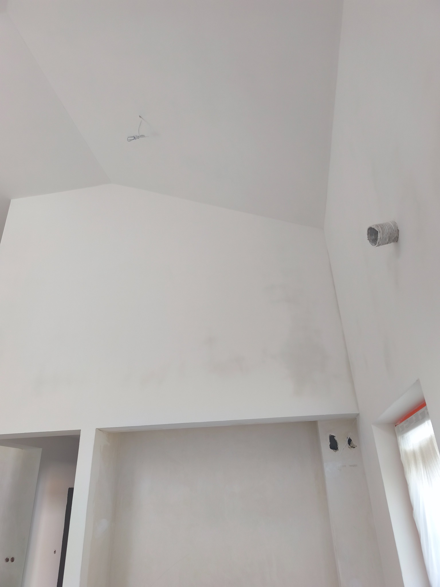 Wnętrze po szpachlowaniu i malowaniu na biało. Widoczna wysoka ściana, sufit ze skosem oraz niedokończone gniazdka elektryczne. Na ścianie widoczny przewód wentylacyjny.