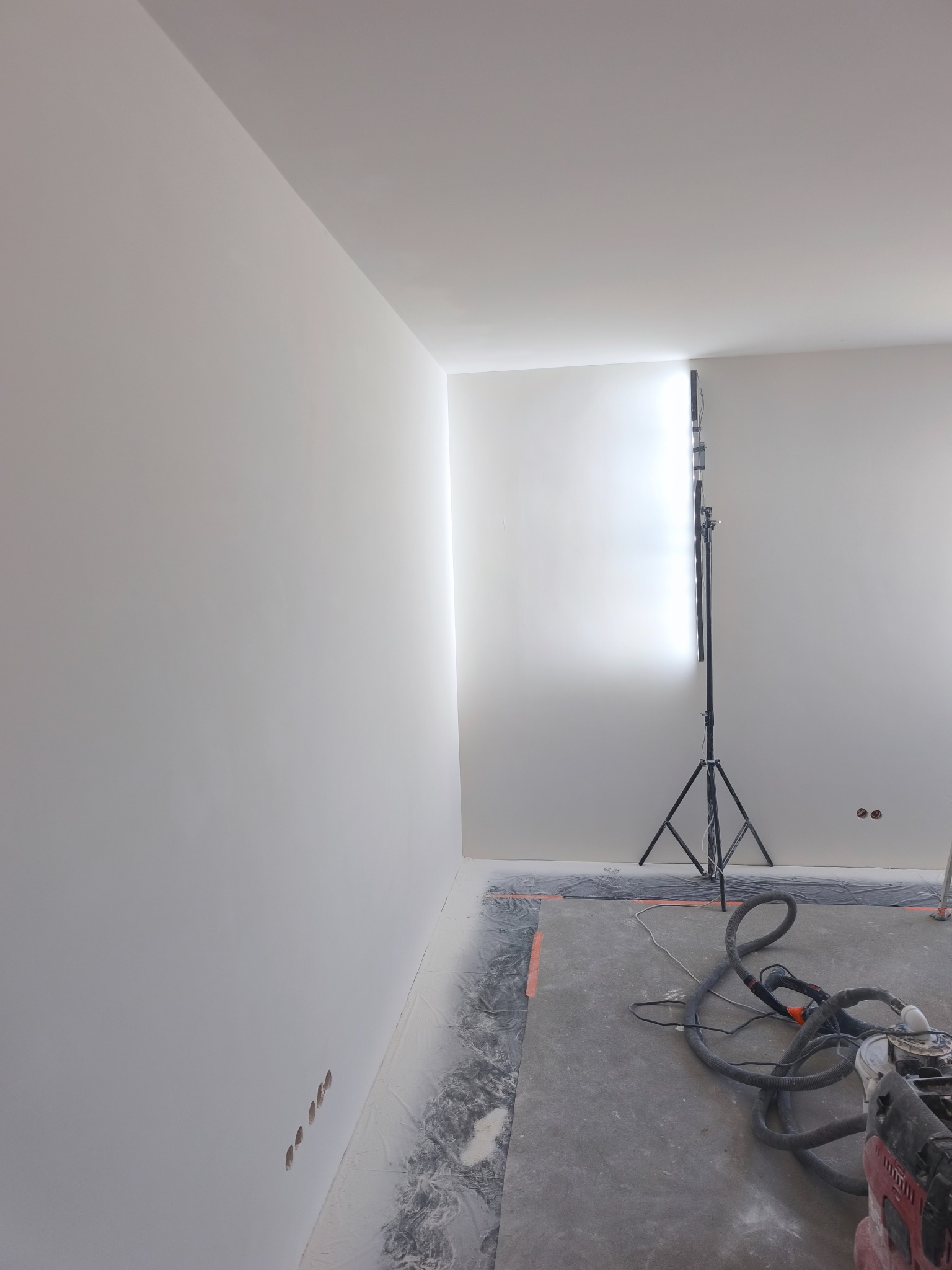 Wnętrze po szpachlowaniu: gładkie, białe ściany podświetlone lampą budowlaną. Na podłodze widoczny sprzęt do szlifowania i zabezpieczenia.