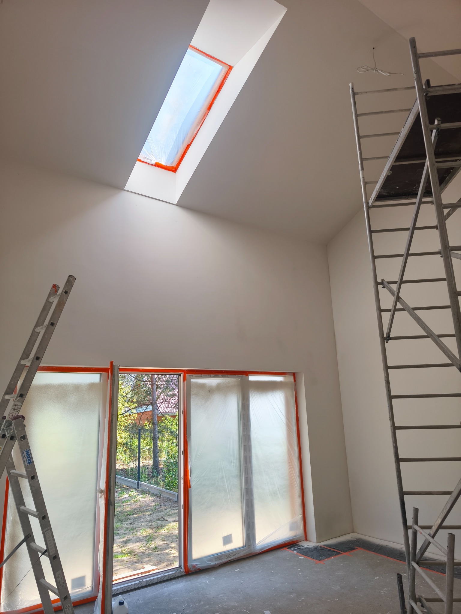 Białe ściany w pokoju ze skosami i oknem dachowym zabezpieczonym folią. Widoczne drabiny, podłoga zabezpieczona folią malarską. Przygotowanie do malowania.