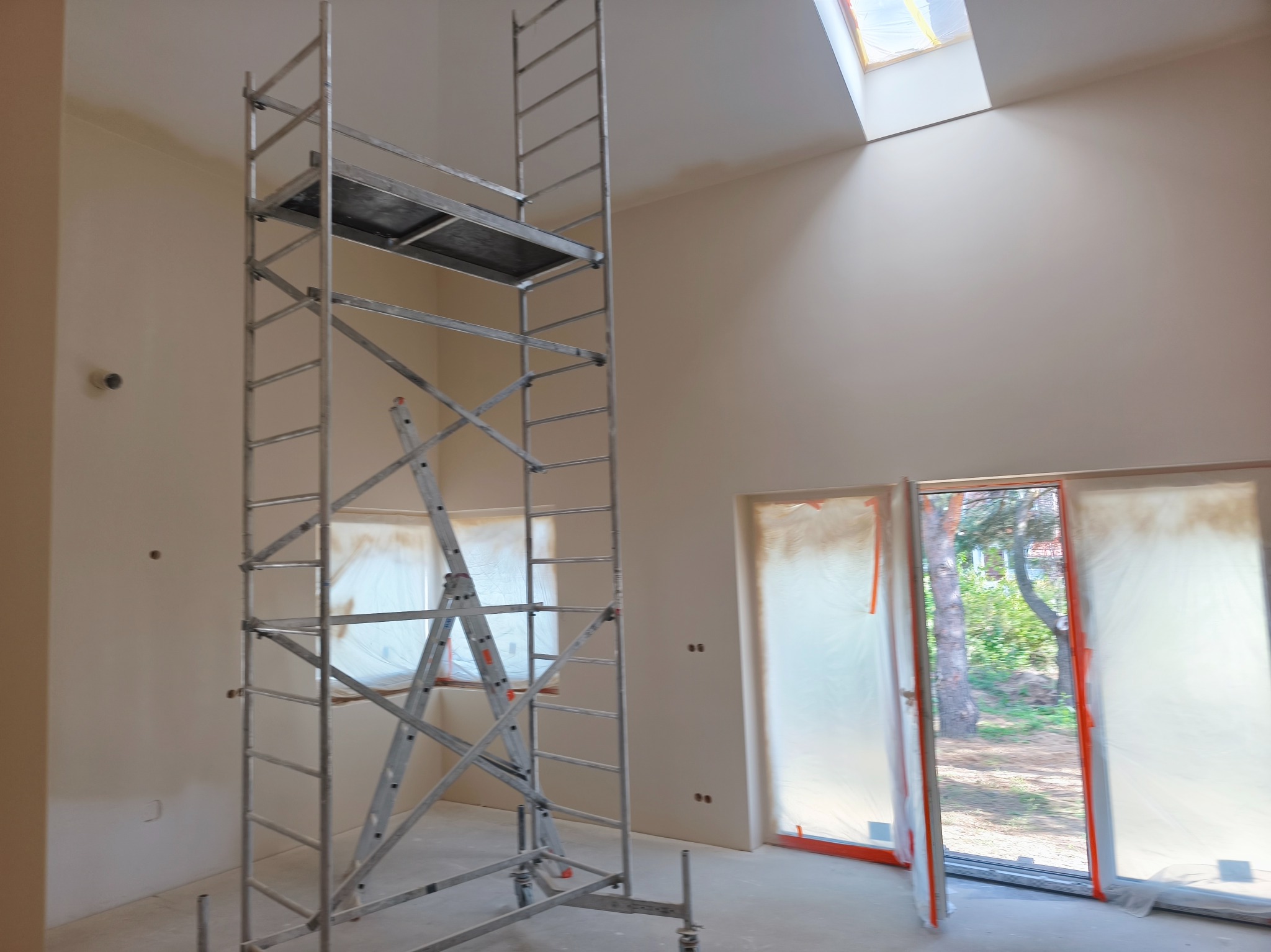 Wnętrze w trakcie prac wykończeniowych: ściany w jasnym odcieniu, rusztowanie na kółkach, okna zabezpieczone folią malarską, widoczne okno dachowe.