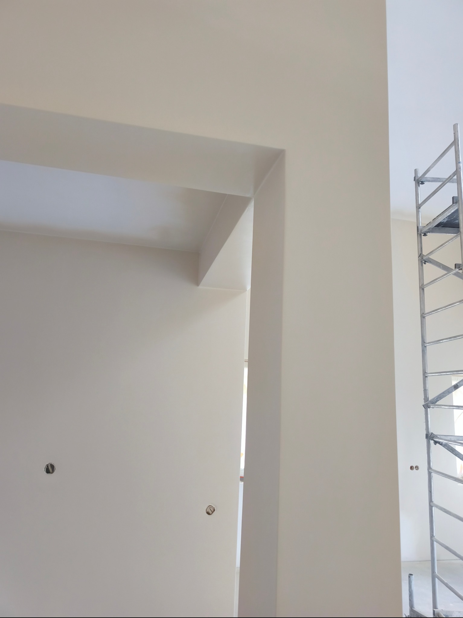 Świeżo pomalowane na beżowo ściany z widocznymi otworami na gniazdka i fragment rusztowania, ukazujące etap wykończeniowy w jasnym wnętrzu.