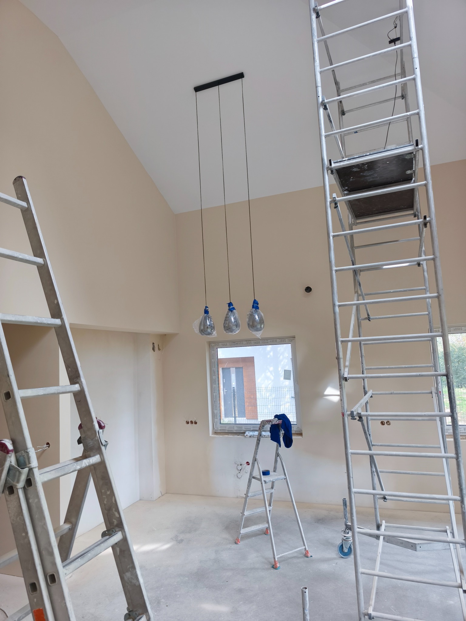 Wnętrze w trakcie malowania: ściany w odcieniach beżu, drabiny malarskie, aluminiowy ruszt, wiszące lampy z żarówkami w folii ochronnej przy oknie.