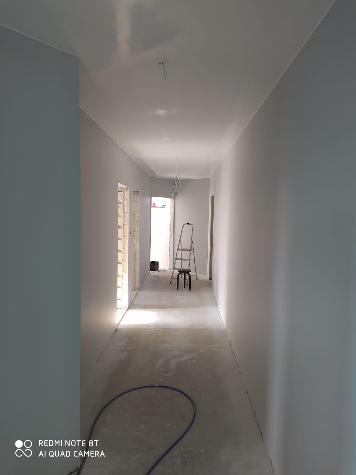 Świeżo pomalowany korytarz w odcieniach szarości i błękitu, z widocznymi śladami gładzi na suficie, drabina malarska, krzesło i zwisające kable elektryczne, podłoga w stanie surowym.