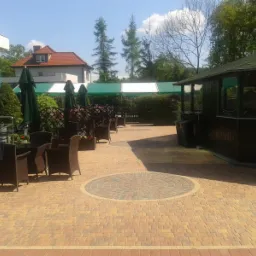 Dziedziniec z kostki brukowej w odcieniach beżu i brązu, z okrągłym wzorem, otoczony meblami ogrodowymi z rattanu, zielonymi parasolami i roślinnością, budynki w tle.