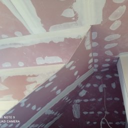 ANDYMAL firma remontowa Andrzej Firmuga - Skośny sufit poddasza w trakcie szpachlowania, widoczne nierównomiernie nałożone białe plamy gładzi na różowym podkładzie, krawędzie zabezpieczone taśmą, widoczne przewody.