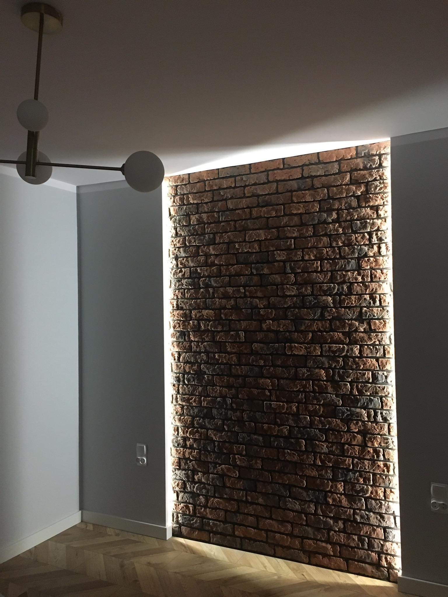 Ściana z cegły klinkierowej z podświetleniem LED tworzącym pionową szczelinę światła, widoczny fragment drewnianej podłogi w jodełkę i nowoczesna lampa sufitowa z kulistymi kloszami.