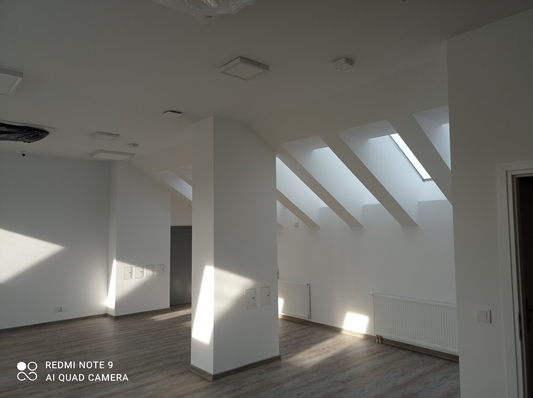 Wnętrze biura po remoncie z białymi ścianami, drewnopodobną podłogą, grzejnikami i oknami dachowymi w skosach, oświetlone silnym światłem słonecznym.