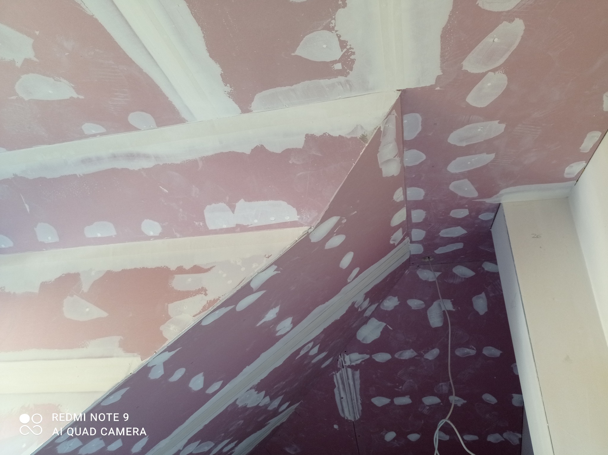 Skośny sufit poddasza w trakcie szpachlowania, widoczne nierównomiernie nałożone białe plamy gładzi na różowym podkładzie, krawędzie zabezpieczone taśmą, widoczne przewody.