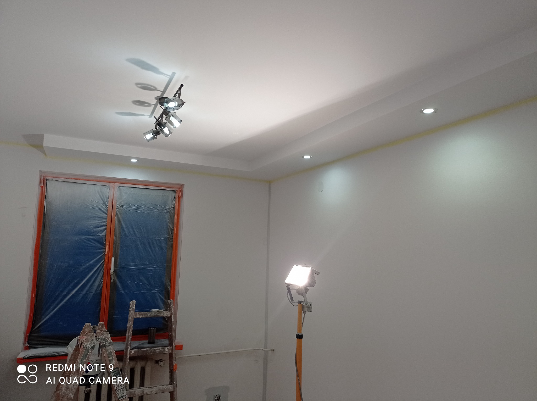 Pokój w trakcie odświeżania: okno zabezpieczone folią, ściany świeżo pomalowane na biało, widoczna drabina, lampa halogenowa na statywie oświetlająca ścianę, taśma malarska przy suficie.